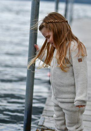 Mono de forro polar gris con mangas largas, cremallera lateral y un parche con el logo en la manga; modelo con cabello rubio ondulado en una trenza.