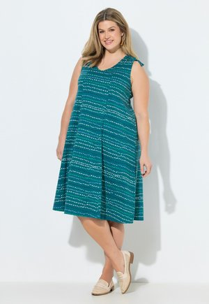 Uśmiechnięta kobieta w turkusowej, bezrękawowej sukience do kolan z białymi kropkowanymi paskami i beżowymi plecionymi mokasynami, stojąca na białym tle.