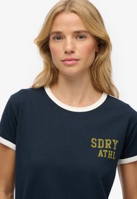 Marine t-shirt met crèmekleurige ringerhals en mouwen, met daarop een groene logo-afdruk "SDRY ATHL" aan de bovenkant van de linkerkant.