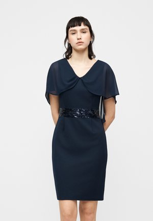 Γυναίκα με κοντά σκούρα μαλλιά, που φοράει μπλε φορέμα μήκους μέχρι το γόνατο με διάφανα μανίκια μανδύα και ζώνη με παγιέτες, στέκεται μπροστά σε λευκό φόντο.