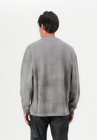 Felpa grigia con finitura testurizzata e usurata; scollatura e polsini a costine; vestibilità rilassata; indossata con pantaloni neri.