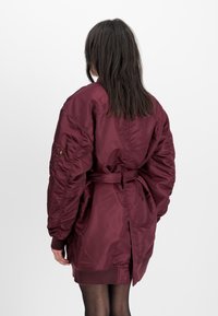 Maroon puffer jacket with a belted waist, ribbed cuffs, and a vented back. Made from shiny, smooth fabric with a loose fit.