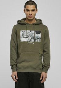 Oliven grønn hettegenser laget av myk tekstil, med et grafisk motiv av en tatovert hånd som holder kjeder og ordet "pray" i hvit tekst på forsiden.