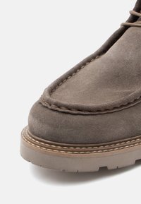 Botín de ante marrón hasta el tobillo con punta redondeada, costuras visibles y suela de goma texturizada que presenta un borde de color claro en contraste.