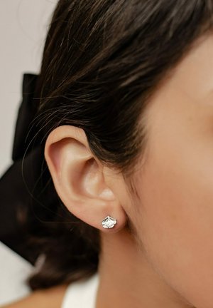 Silberne Steckerohrringe in Blattform mit strukturierten Details, vor glattem, dunklem Haar und einem neutralen Hintergrund.