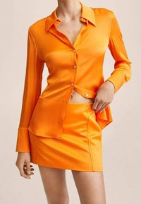 Blusa di seta arancione brillante con design colletto, chiusura con bottoni e maniche lunghe, abbinata a una mini gonna coordinata. Texture liscia in tutto il capo.