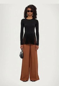 Sieviete ar cirtainiem matiem, valkājot melnas saulesbrilles, melnu garu piedurkņu topu ar frizi, platas brūnas bikses, tur rokā mazu melnu roku somu.