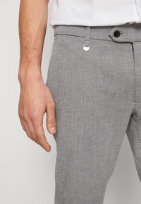 Šedé a černé kárované kalhoty s hladkou texturou, které mají přední kapsu a malý kovový akcent na pásku.