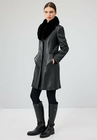 Czarny skórzany płaszcz z futrzanym kołnierzem, o dopasowanym kroju, z zapięciem na guziki i bocznymi kieszeniami. Nosi się go na czarne legginsy i wysokie kozaki.