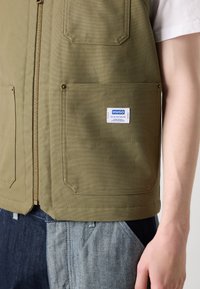 Olive green vest with a front zipper, two large pockets, and a brand label. Made from durable fabric, features a textured finish.