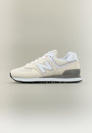 Baskets New Balance beige et blanches avec dessus en mesh et daim, design à lacets et semelle grise rembourrée sur fond beige.