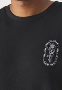 Persona que lleva una camiseta negra con un gráfico blanco de una mano esquelética sosteniendo una rosa, rodeada de texto circular en el área del pecho izquierdo.