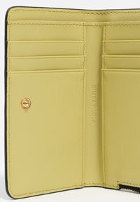 Interno di un portafoglio di pelle gialla con più slot per carte, accenti neri, bottone in metallo dorato e marchio impresso sul lato.