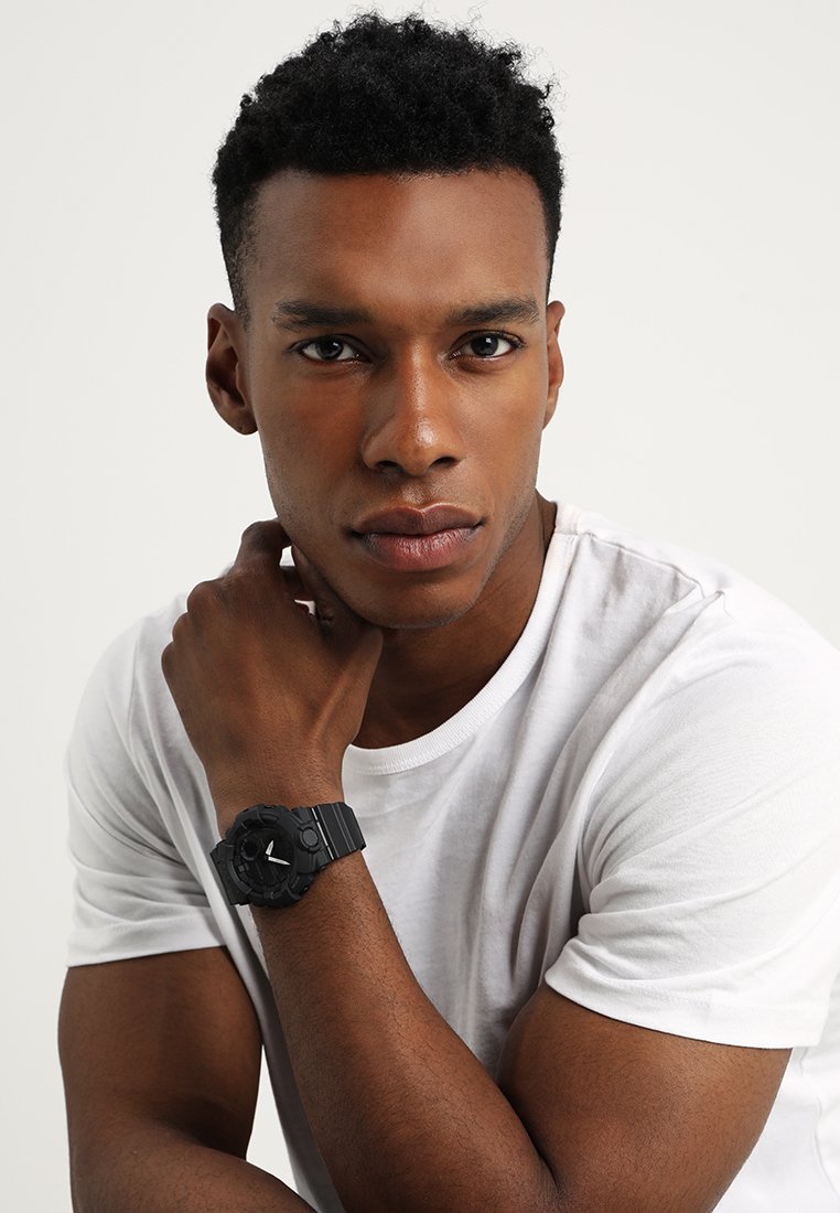 Montre de sport noire avec un bracelet texturé, un cadran rond et des marqueurs d'heure blancs, portée sur un poignet nu sur un fond de chemise blanche unie.