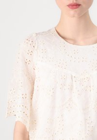 Vit broderad blus med korta ärmar, med blommönster, texturerad tyg och rund halsringning. Fint stygn över hela.