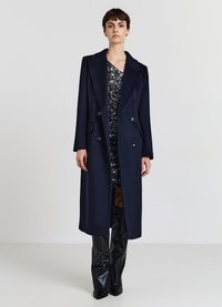 Cappotto doppiopetto in misto lana blu navy con grandi bottoni, ampie rever e tasche laterali, indossato sopra un abito nero scintillante e pantaloni in pelle.