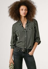 Camicia a quadri bianca e nera con colletto, maniche arrotolate e tasche sul petto, abbinata a jeans scuri e una cintura decorativa.