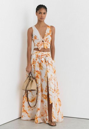 Femme portant une tenue deux pièces florale blanche et orange avec un crop top noué, une jupe taille haute fendue, tenant un sac à main beige.