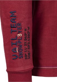 Maglietta bordeaux a maniche lunghe con testo ricamato: "VAIL TEAM" e "SNOWPLAYER" in blu, con coordinate in rosso e crema sulla manica.
