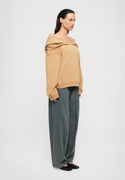 Femme se tenant de côté, portant un sweat à capuche beige surdimensionné, un jean large gris délavé et des chaussures noires à enfiler sur un fond blanc.