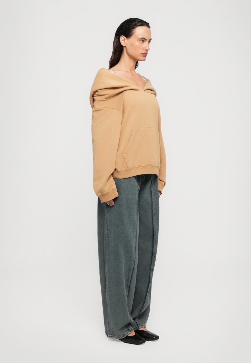 Kvinna som står på sidan iklädd en överdimensionerad tan off-shoulder hoodie, utsvängda grå jeans och svarta slip-on skor mot en vit bakgrund.