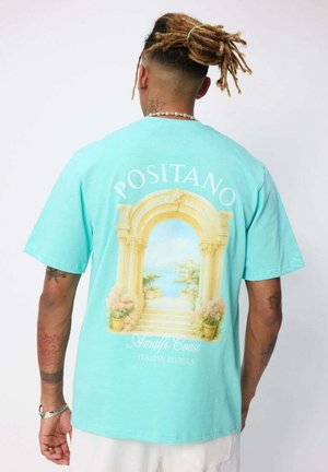 Homme aux dreadlocks vu de dos, portant un T-shirt turquoise avec une impression de l'arche de Positano et une scène côtière au dos.