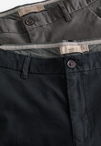 Zwei Paar taillierte Hosen in Dunkelgrau und Schwarz, mit Knopfverschlüssen und Etiketten. Der Stoff wirkt glatt mit einem leichten Glanz.