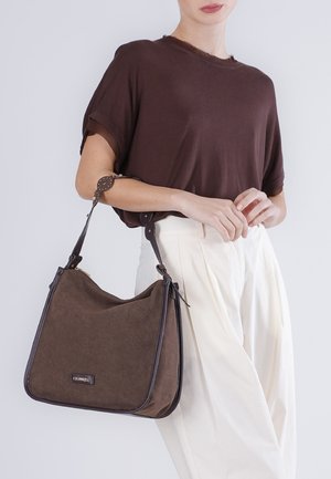 Mujer con una blusa marrón y pantalones blancos, sosteniendo un bolso de hombro de ante marrón con correas de cuero y una etiqueta metálica con el logo.