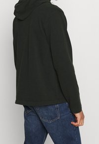 Dark green hoodie with a soft texture and hood, featuring long sleeves and a relaxed fit, paired with blue denim jeans.