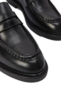 Mocassins en cuir noir avec des détails de couture, présentant une finition lisse et un nez rond, montés sur une semelle en caoutchouc à faible profil.