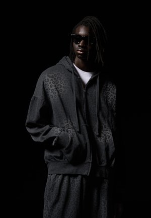 Mladá osoba s dredmi, ktorá nosí čierne slnečné okuliare, tmavosivú mikinu so vzorom leopardej kože a zodpovedajúce nohavice na čiernom pozadí.