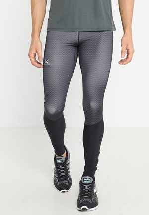 Homme portant un legging de sport noir et gris à motifs et des chaussures de course noires, debout sur un sol blanc.
