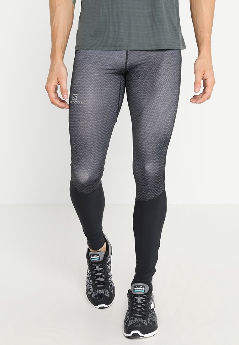Homme portant un legging de sport noir et gris à motifs et des chaussures de course noires, debout sur un sol blanc.