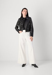 Een vrouw staat zelfverzekerd, gekleed in een zwart leren jasje, een zwart topje, witte wijde broek en zwarte puntige hakken, tegen een lichte achtergrond.