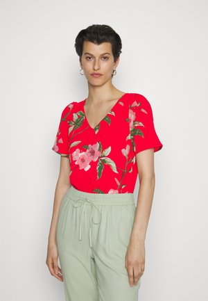 Camisola curta vermelha de flores com decote em V, apresentando flores cor-de-rosa e folhas verdes, combinada com calças verdes claras com cordão.