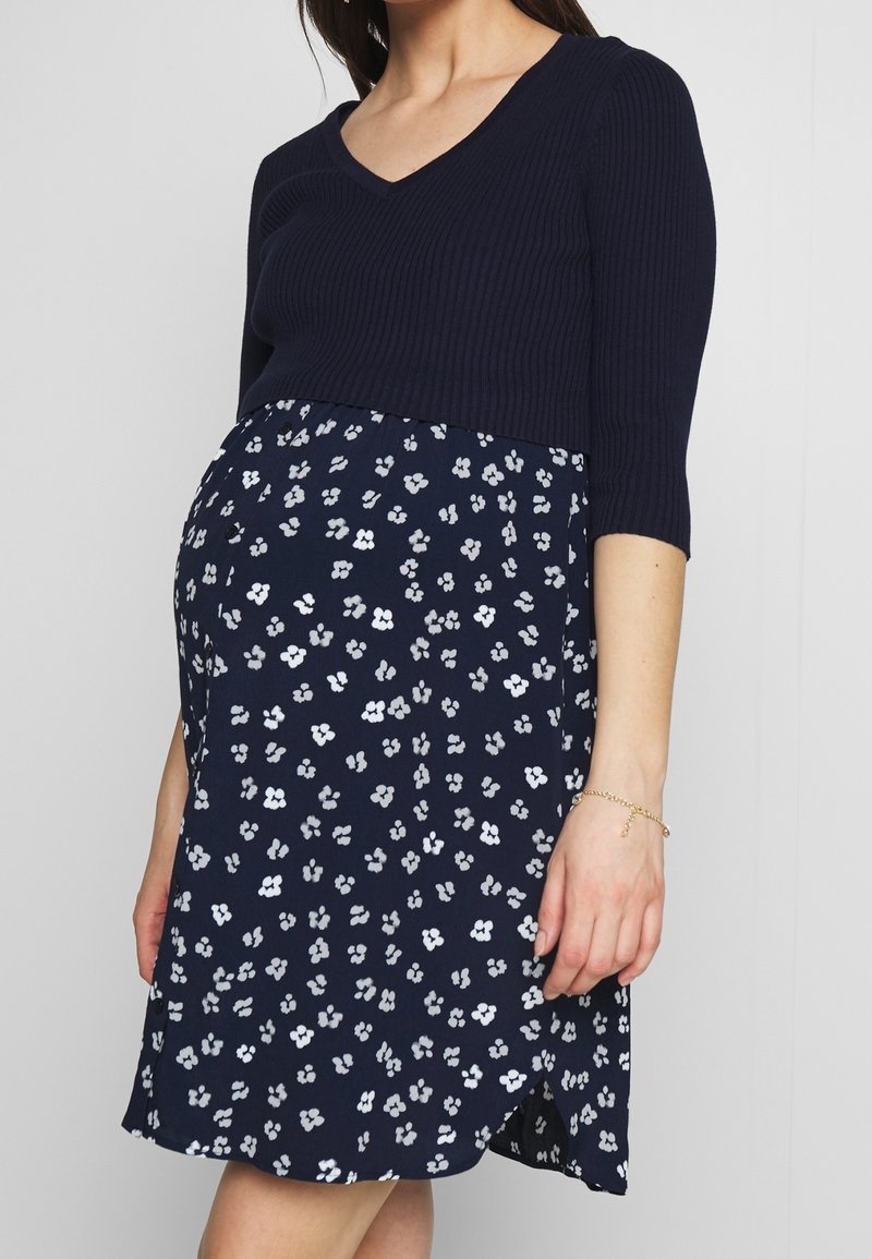 Femme enceinte portant une robe bleu marine à motifs floraux blancs et un haut côtelé bleu marine à manches trois-quarts.