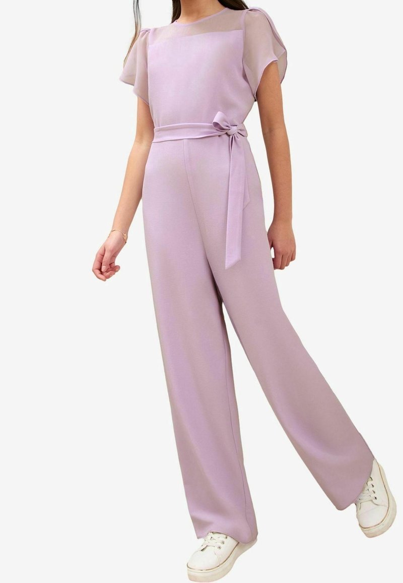 Femme portant une combinaison pantalon ample violet clair avec un empiècement transparent, une ceinture à nœud et des baskets blanches, debout devant un fond uni.
