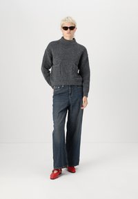 Grijze gebreide trui met een opstaande kraag en voorzakken, gecombineerd met losse donkere spijkerbroek en rode platte schoenen.
