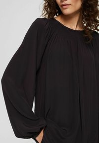 Haut noir à manches longues avec une coupe décontractée, détail de col smocké et tissu drapé en douceur. Comprend un léger éclat et des manches amples.