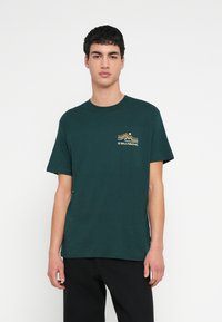 Tmavozelené bavlnené tričko s krátkym rukávom a okrúhlym výstrihom. Na ľavej strane na hrudi má malý viacfarebný vlnený logo.