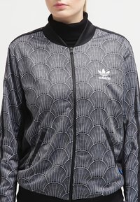 Černobílé zipové sako s vlnkovaným vzorem, hladkou texturou a černými akcenty, obsahující boční kapsy a žebrovaný límec.