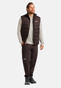 Zwarte gewatteerde vest met hoge kraag, gedragen over een lichtgrijs langetermijnshirt. Gecombineerd met bruine broek en wandelschoenen. Eenvoudige branding zichtbaar.