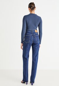 Blå denimjeans med hög midja och raka ben, med två bakfickor och en beige lädermärkespaste. Tyget ser stadigt ut.