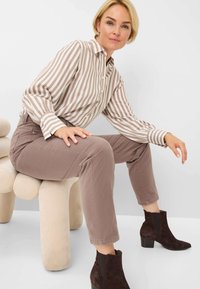 Gestreepte blouse met lange mouwen in beige en bruin, gecombineerd met lichtbruine broeken en donkerbruine enkellaarsjes met een stevige hak, zittend op een zachte, ronde kruk.