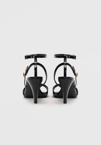 Par sorte svarte patent lær høyhælte sandaler med ankelremmer og gullspenner, sett bakfra mot en hvit bakgrunn.