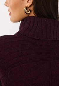 Maglione a maglia bordeaux con colletto alto a coste e dettagli testurizzati. Indossato con un grande orecchino a cerchio d'oro posizionato all'orecchio sinistro.