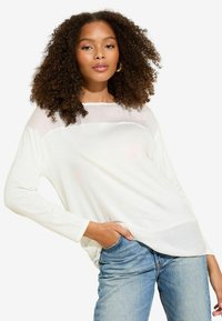 Top bianco a maniche lunghe con pannello trasparente sul petto, abbinato a jeans denim azzurri. Modello presenta una vestibilità ampia e scollatura rotonda.
