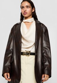 Blazer oversized din piele maro peste un top alb drapat, asortat cu o curea neagră cu cataramă circulară aurie. Fustă bej cu nasturi.
