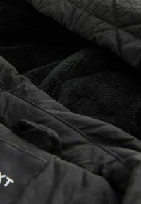 Veste matelassée noire partiellement ouverte pour révéler une doublure polaire noire douce à l'intérieur, montrant un contraste dans les textures des tissus.