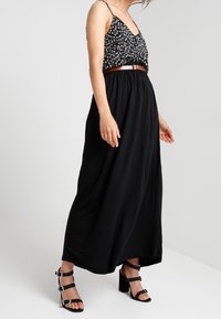 Svart maxiklänning med en åtsittande överdel, dekorativt blommönster och justerbara band, kombinerad med svarta blockklackssandaler med nitar.